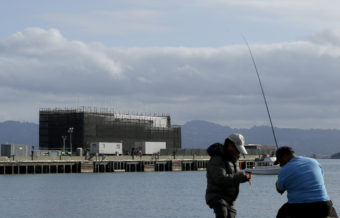 Two men fish in the water in front of a barge on Treasure Island in San Francisco on Tuesday. An unnamed source tells CBS the barge carries a building "constructed of interchangeable 40-foot shipping containers that can be assembled and disassembled at will, allowing it to be placed on barges, trucks or rail cars and taken anywhere in the world." Jeff Chiu/AP