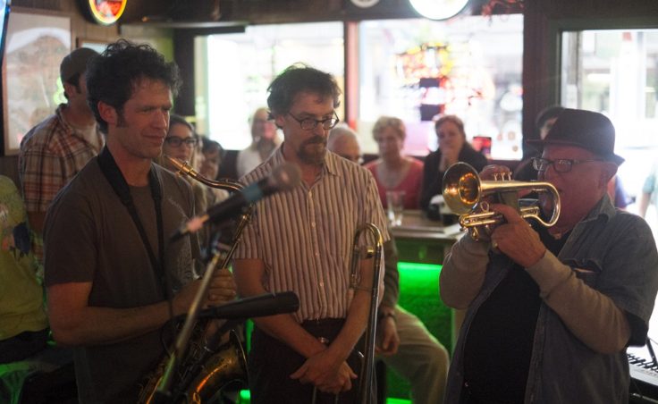 A man plays a cornet while two other men watch him in a bar