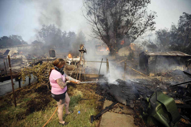 A woman douses debris around her home as her neighbor's home burns Thursday in Escondido. One of the nine wildfires burning in San Diego County suddenly flared Thursday afternoon and burned close to homes, triggering thousands of new evacuation orders. Gregory Bull/AP