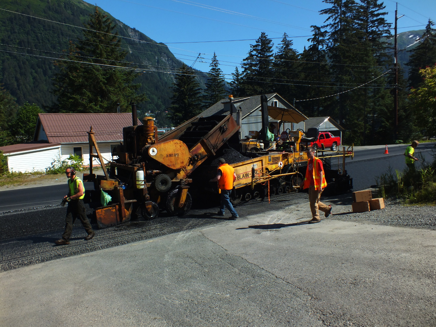 An asphalt spreader follows the long pile of asphalt mix laid down by the tractor trailer. Speed of the spreader sometimes determines the width, height and crowning of asphalt layer. (Photo by Matt Miller/KTOO)