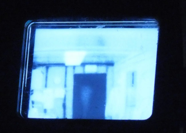 Thermal Imaging Camera or TIC can be used to detect a heat source hidden inside walls or find a victim in a dark, smoky room. Cooler objects, like the exterior door shown in the center of this viewfinder's image, appear as dark or black. Warmer objects, such as the recently turned off ceiling light fixture, show up as bright white. (Photo by Matt Miller/KTOO)
