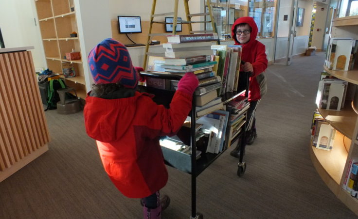 Sadie Frances Curtis, 5, and Q Curtis, 8, wheel in a cart of 119 books they checked out from the old Mendenhall Mall Library just before it closed. Along with their mom Linda Curtis, they were deemed Checkout Champions as patrons who helped with moving books from the old library. (Photo by Matt Miller/KTOO)