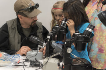 Seven-year-old Anna Nukapigak looks through a microscope at samples. Jeff Rasic and the rest of the students look on.