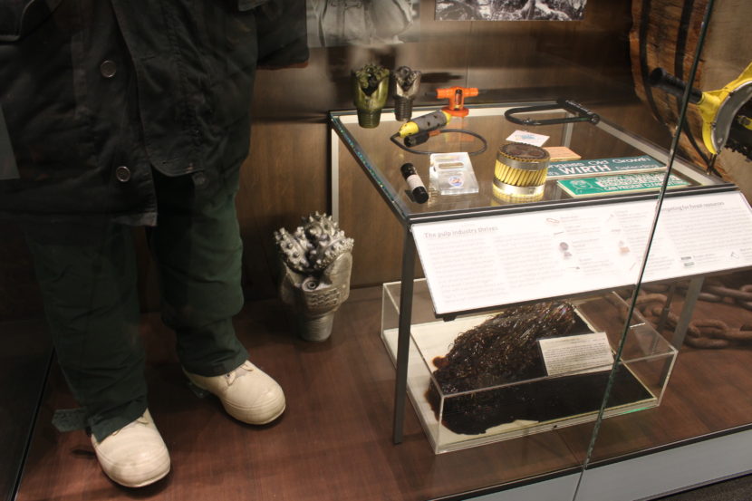 Inside a display case, clothing worn during the construction of the Trans Alaska pipeline, a drill bit, and a soaked pom pom from the Exxon Valdez oil spill. (Photo by Elizabeth Jenkins/KTOO)