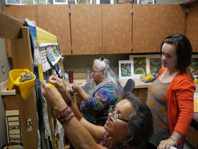 Della Cheney, left, and Clarissa Rizal work on braiding the side borders for the Weavers Across Waters Chilkat/Ravenstail community robe on Monday, August 22, 2016, at the Sealaska Heritage Institute, Juneau. One of three of Rizal's daughters, Lily Hope watches the weavers work on the robe, which will be part of the Huna Tribal House opening celebration. Hope is also an accomplished weaver, actress and storyteller. (Photo by Tripp J Crouse/KTOO)