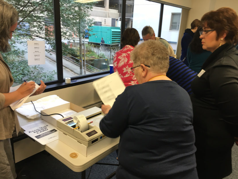 An election official feeds ballots into an optical scanner, while observers watch. Sept. 12, 2016. (By Andrew Kitchenman/KTOO)