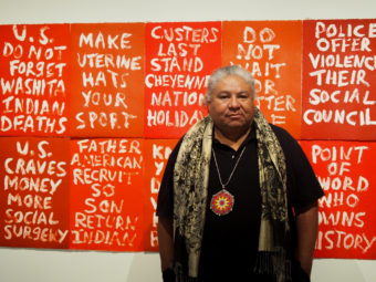 Artist Edgar Heap of Birds stands before his piece “Dead Indian Stories” on display in the “Without Boundaries” exhibit. (Photo by Zachariah Hughes/Alaska Public Media)