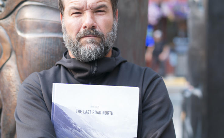 A man in front of a totem holds a book. Juneau photographer Ben Huff holds his book 'The Last Road North' following the demonstration against budget cuts to the Alaska State Council on the Arts. (Photo by Annie Bartholomew/KTOO)