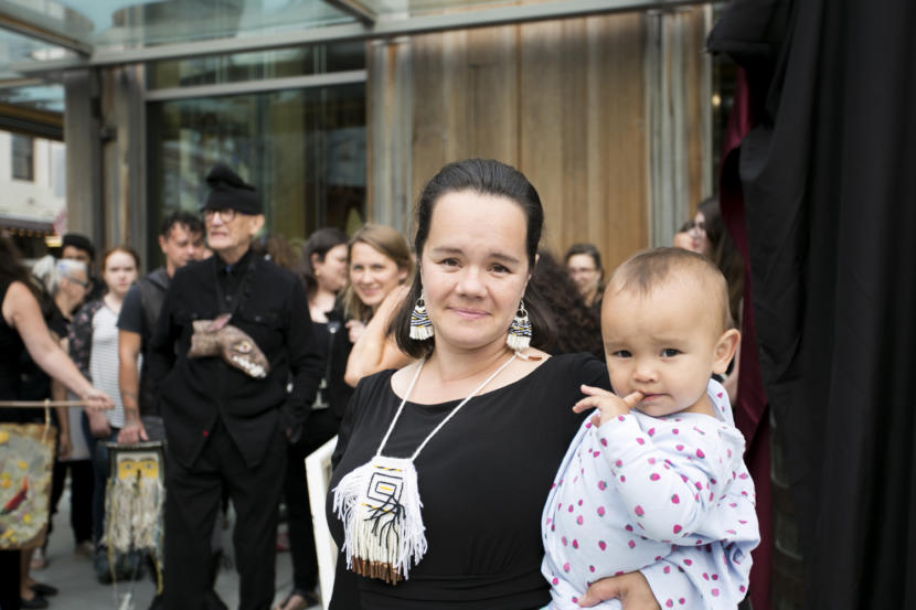 A woman holds her baby on a street corner in Juneau. Master Chilkat Weaver Lily Hope holds her daughter at the peaceful demonstration opposing the budget cuts to the Alaska State Council on the Arts outside Sealaska Heritage Institute on Tuesday, July 9, 2019. Hope helped organize the event which covered the totems with black fabric to create a public art "black out." Photo by Annie Bartholomew/KTOO.