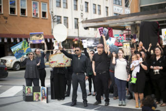 People dressed in black hold up paintings, books, drums, and artwork on a street corner in Juneau. Juneau artists hold up their work at a demonstration outside Sealaska Heritage Institute protesting the budget cuts to the Alaska State Council on the Arts on Tuesday, July 9, 2019. (Photo by Annie Bartholomew/KTOO)