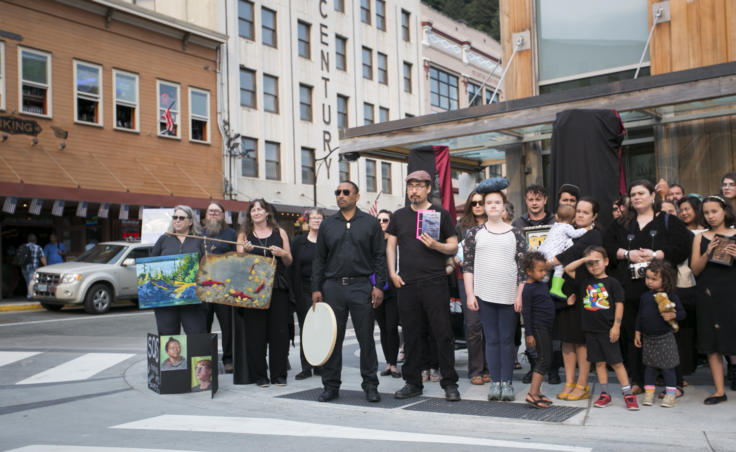 A photo of people holding books, painting, drums and art on a street corner. They are dressed in black.