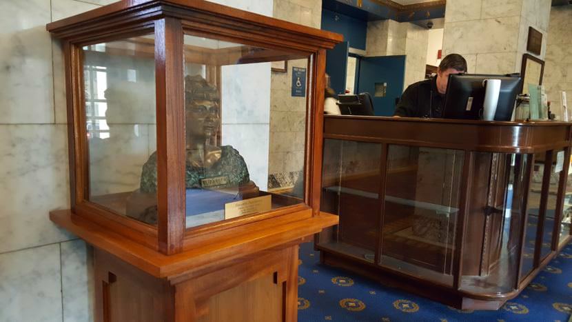 Ryan Strickland, a security and information specialist for the State of Alaska, works the front desk of the Alaska Capitol on Tuesday, July 11, 2017, in downtown Juneau. In the front lobby a bust of Alaska Native civil rights leader Elizabeth Peratrovich greets capitol visitors. (Photo by Tripp J Crouse/KTOO)