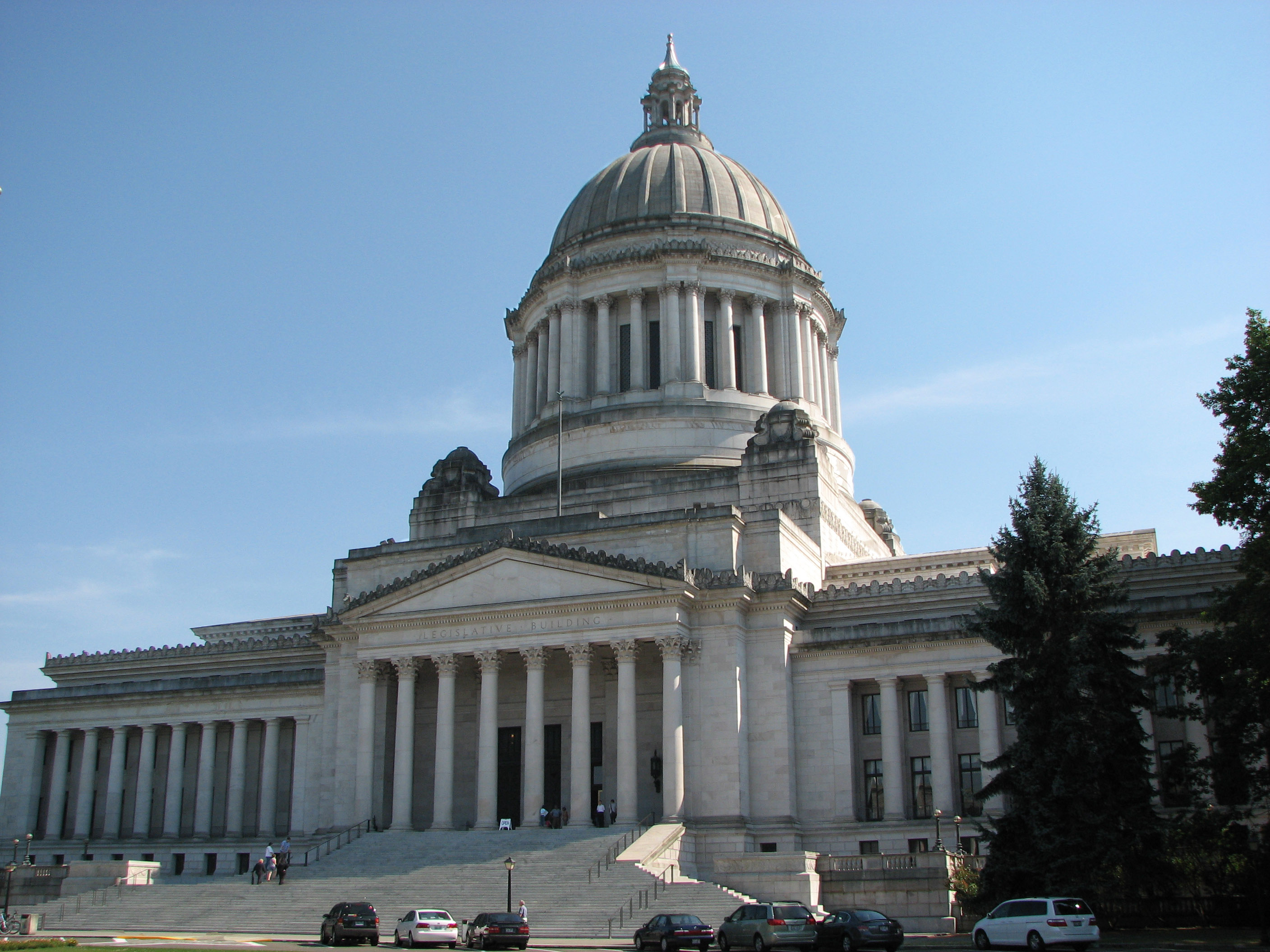 The Legislative Building of the Washington State Capitol, Olympia. The building is in afternoon sun. An "open" sign invites visitors inside to see the dome interior, tiffany chandelier, marble panels, and artwork. (Creative Commons photo by Gerald Hawkins)