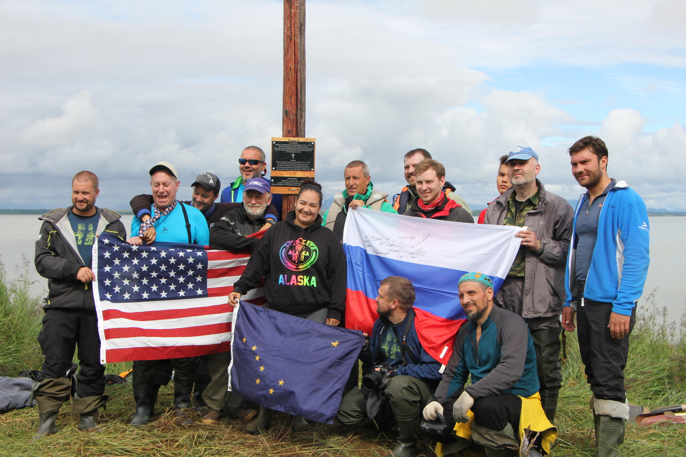 The Russian team and the Dillingham residents who helped to place the monument at Nushagak pose with the American, Russian, and Alaskan flags. (Photo by Allison Mollenkam/KDLG)