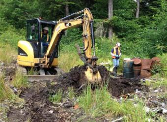 A backhoe digs up part of the old Byford Junkyard in Wrangell in 2014. After removing old cars, oil drums and other trash, the state is treating and moving contaminated soil to a rock quarry south of town. (Photo courtesy Department of Environmental Conservation)