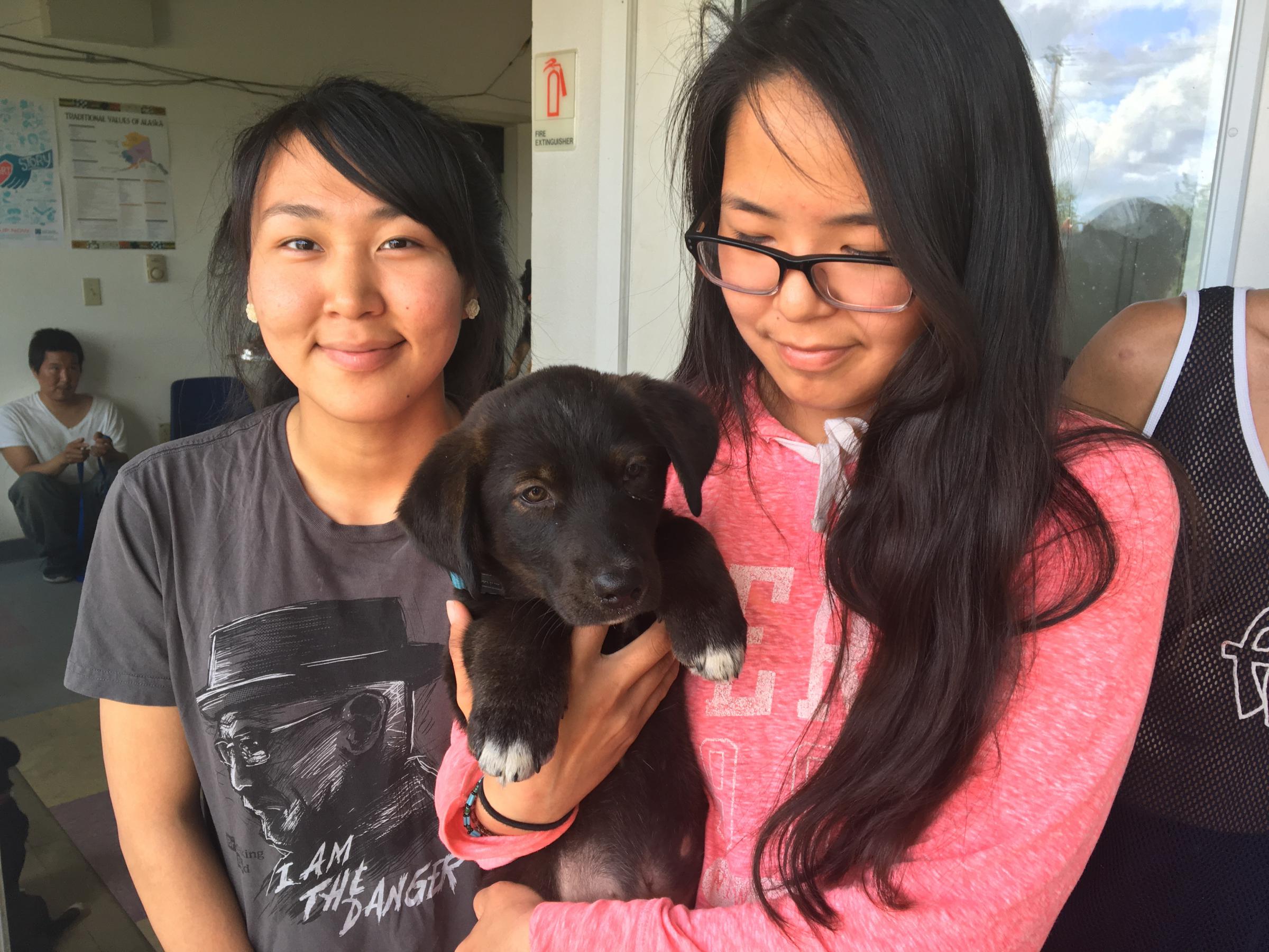 Two girls in Kwethluk wait with a puppy at the local fire station, where three vets from the non-profit Alaskia Native Rural Veterinary were working for the day. (Photo by Teresa Cotsirilos/KYUK)