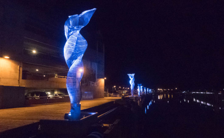 Nighttime, looking down a row of 20 foot tall metal sculptures mounted on the edge of a dock and lit up with bright blue light. The sculptures are made of many thin strands of metal swirling around a central core from the base to the top.