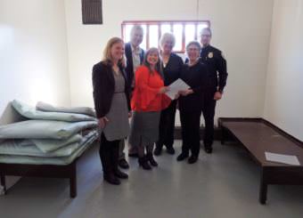 Heather Parker with Gov. Walker’s office, DOC Commissioner Dean Williams, Pretrial Director Geri Fox, Haines Mayor Jan Hill, Haines Borough Manager Debra Schnabel and Haines Police Chief Heath Scott pose in a Haines jail cell with a revised community jails contract. (Photo by Emily Files/KHNS)