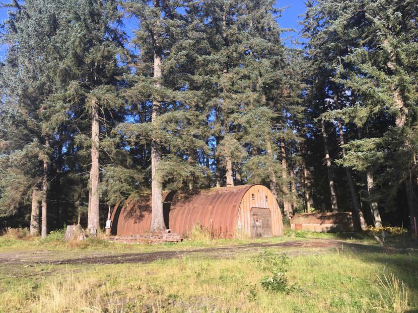 Two Quonset huts at the corner of Mendenhall Loop Road and Atlin Drive are among the last remnants of Juneau’s history during World War II. Skip Gray, a crew chief for Gavel Alaska, asked about the huts’ history for this edition of Curious Juneau. If you’ve driven on Mendenhall Loop Road, you may have seen the huts. The rusted steel half-circles look a bit like mini-airplane hangars. I didn’t know much about Quonset huts, so I called Julie Decker. She’s the director of the Anchorage Museum – and co-edited the book “Quonset Hut: Metal Living for a Modern Age.” Decker described a Quonset hut as “a temporary structure, born of war and expediency, and ended up becoming a permanent fixture on our landscape.” The huts were named after Quonset Point, Rhode Island, near where they were first built at a naval air station. The military developed them at the start of World War II as solid, inexpensive structures that were easy to ship and build. They were used around the world as places for soldiers to eat, sleep, work, and heal. Decker said that after the war, the military and local governments, including Anchorage, had to decide what to do with them. Many were sold off. “They tried everything from banning Quonset huts from our downtown to using them for railroad housing,” she said. “And, instead, they became permanent fixtures and got used as artists’ studios and garden spaces and sheds and restaurants. And I just kind of loved the way these things got adopted and became part of the fabric of our lives.” They remained more common in Alaska than they were in other places after the war. In part, that’s because it was more expensive to remove them here. So people had to find ways to use them. Many of the modest structures became houses. I told Decker about one in Juneau’s quirky Starr Hill neighborhood. It’s currently listed at $179,000. Decker asked if I was kidding. I wondered what it was like to live in one. It doesn’t look like the Quonset huts on Atlin Drive have been lived in for a long time. But a coworker let me know that North Douglas resident Paul Disdier still lives in one, so I stopped by his house to find out what it was like. Disdier said: “The roof that was on this house, the steel was so thick, you actually wouldn’t even need to paint it. It would still be here. You know, I think the ones that are out in the valley have the original roofs on them and they’re unpainted still. It’s so thick it would take forever to rust through them.” There are odd parts to living in the structure. The metal roof can make it damp. But one problem for today’s residents wouldn’t have affected the soldiers in the war. “This house has a lot of steel in it,” he said. “I have not very good cellphone reception when I’m in the house and that’s just what you have to live with.” At this point, I knew about the Quonset huts in other parts of Juneau, but I was stuck on Atlin Drive huts. I went back to Skip Gray for help. “I had heard stories of Quonset huts in the Mendenhall Valley much of my life it seems like, off and on, just various people talking about them,” he said. “I think I’d noticed them in the woods before, but they had become much more visible, because some trees have been cut down around them. … I knew they were related to the military somehow, but I didn’t know how.” Skip gave me a lead that led to Mary Lou Spartz. She’s a spry 86-year-old who has vivid members of life in Juneau during World War II. “It was intended to be a big base,” Spartz said. “And it was camouflaged from the road, so we never saw any of it.” Like other civilians, Mary Lou wasn’t allowed on the base. But she remembered there was a full hospital on it. “The thinking was, (if) there was a major invasion of Alaska, and there were wounded, and people had to be brought somewhere, they would come here for medical attention,” she said. Spartz said Juneau residents were worried that the Japanese military would invade the city. “We just didn’t have any idea,” she said. “As you understand, we’re close enough that it wouldn’t have been that hard.” Her father made concrete using a gravel pit he owned that was behind where the Quonset huts stand today. “He was pretty much put out of business by the Army,” Spartz said. “They were going to expand the airport. And they just gave him a price that was way under what it was valued, and said take or leave it, but we’re going to take it.” She recalled that the base had many Quonset huts, which were sold and spread around town after the war. “They were substantial,” Spartz said. “If you look at the Quonset hut that is out there at Atlin, you can tell that it’s meant to last.” I stopped by the Juneau-Douglas City Museum to see if they had information on the huts. The staff found a description of the Army post written during the war by its commanding officer, Lt. Col. Roy Riegle. You get a sense of his proper personality from his report. He proudly writes that the post didn’t have a single case of venereal disease. When the soldiers left their Quonset huts, Riegle ordered them not to visit the legal brothels on South Franklin Street. The Army post covered 500 acres along what is now Mendenhall Loop Road. It housed 2,000 soldiers – more than a third of the city’s entire population in the census taken the year before the war. The post was self-sustaining, with its own laundry, shoe shop and bakery. A farm along Eagle River plowed by the soldiers provided potatoes, radishes, lettuce and cabbages. Many of the soldiers’ jobs were to guard the airport, which was under construction immediately before the war. It served as a link between the Lower 48 and air bases in western Alaska. The post also provided basic training to soldiers drafted from Southeast Alaska. Riegle mentions the post’s Quonset huts, saying that their dark interiors were painted white to make them appear brighter. I didn't have to go far to find more. Next door from KTOO, Anastasia Tarmann, a librarian at the Alaska State Library, remembered an oral history interview she did with Gus Mohr, who died in 2016. Mohr was a sergeant who arrived in Juneau about a week after the attack on Pearl Harbor. He and the other soldiers slept in tents the first winter. Then the pieces of the Quonset huts arrived. “We actually built the Quonset huts,” Mohr said. “They had wood floors that were shipped up from the United States.” Mohr said the post wasn’t a bad place to live. “We had a beautiful camp there, well maintained,” Mohr said. “And we built a big kitchen, hot showers and everything. We used diesel oil, fuel for the hot showers and everything like that.” Mohr said the soldiers ate well, including food they got from fishing and hunting. “We weren’t hurting,” he said. “We shot bear. Bear meat’s pretty stinky, but deer is pretty good, so we had all fresh food and everything like that.” I never found information about the use of these exact huts in the war. One source says he heard the larger one served as a laundry. A Juneau Empire story passed along a legend that Bob Hope – the country’s most popular comedian -- performed in it. We do know from Riegle’s account that Hope visited the post. That Empire story said the Forest Service used the huts after the war, to store firewood and as a space to paint signs and benches. Fast forward to 2010. Developer Richard Harris bought the huts. To find out what’s happening in them these days, I called Harris’s office. The person who answered the phone says one’s being used as a paint shop. While the huts may not have much of a future, they’ve had an interesting past. I reported back to Skip Gray. “I think it’s cool that you found out so much about the military complex,” he said. “I’d never heard how big it was, or why they were there, what they were doing there. … I asked if he’d think of this information when he drives past them in the future. He replied: “Yeah, every time I drive past them, it just kind of plants that little question in my mind of what they were all about. And I’m thankful you were able to dig up some great information on it.” Gray said he used to live in a Quonset hut at the top of 6th Street, adding that it was “Cosy. It was a pleasant place to live.”