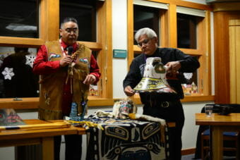 Roy Dennis, left, and Wayne Price tell the story of a boy eaten by sockeye salmon carved into a Tlingit hat. (Photo by Berett Wilber/KHNS)