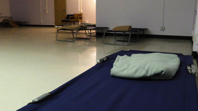 A single dark colored cot with one blanket on it in the foreground, with a couple other cots and some chairs in the background