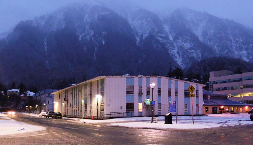 The old Alaska Department of Public Safety Building, a blue and white building, on the edge of Whittier Ave. at dusk with street lights illuminated in front of it and a snow covered mountain visible behind it