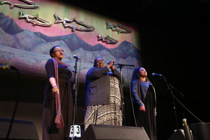 Three women sing in front of a backdrop of metallic fish, the northern lights and a mountain scene. Erika Lee, Jocelyn Miles and Salissa Thole sing as the group "Brown Sugar" at the 44th Annual Alaska Folk Festival in front of the backdrop created by the JDHS Art Club. (Photo by Annie Bartholomew/KTOO)