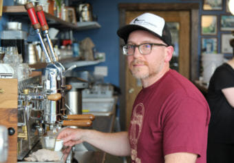 Austin Schwartz, with a short beard, glasses and a baseball cap, holds a cup of coffee under the nozzle of an espresso machine