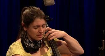A picture of a woman with a bandana on playing cello in a tv studio. Still image of Kat Moore of the Forest That Never Sleeps performing a Red Carpet Concert at KTOO Public Media in April 2018.