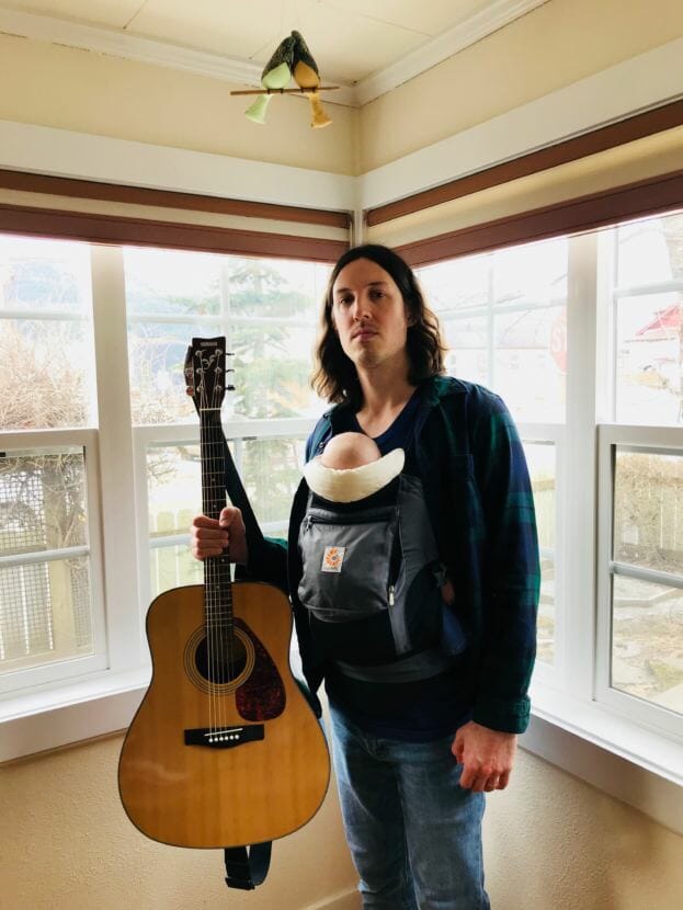 A picture of a man holding a guitar with a baby carrier on his chest. KTOO volunteer DJ Patrick Klacza hosts the radio show Cat Plaza on KXLL 100.7 FM Juneau. (Image courtesy of the artist)