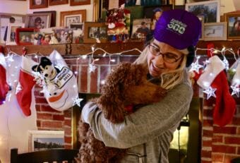 Jackie Klejka wears her signature K300 hat while holding her indoor dog Jersey inside her Bethel home, which is heavily decorated with sled dog paraphernalia. Klejka has been coordinating the K300 veterinary program since 1993. (Photo by Anna Rose MacArthur/KYUK)
