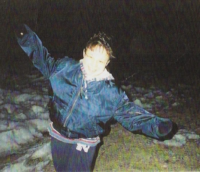 A night photo of a young woman in a blue jacket smiling and playfully holding her arms out from her sides
