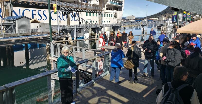 Vicki Logan of Travel Juneau greets and hands out walking maps to passengers of the Ruby Princess at the Franklin Dock on Sunday, April 28, 2019. The Yées Ḵu.Oo dance group performs behind her as part of a welcome party for the first big cruise ship of the season.