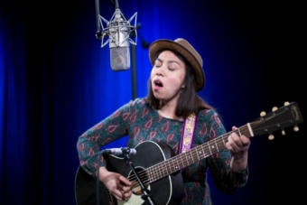 A woman sings into a microphone holding a black guitar