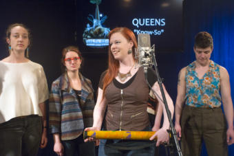 A woman holding a rain stick smiling around a microphone with other women in the background