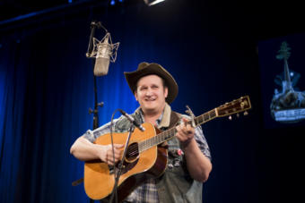A man in a cowboy hat with a guitar sings into a microphone in the KTOO TV Studio in Juneau. Arkansas songwriter Willi Carlisle performs a Red Carpet Concert during the 2019 Alaska Folk Festival in the KTOO TV Studios. (Photo by Annie Bartholomew/KTOO)