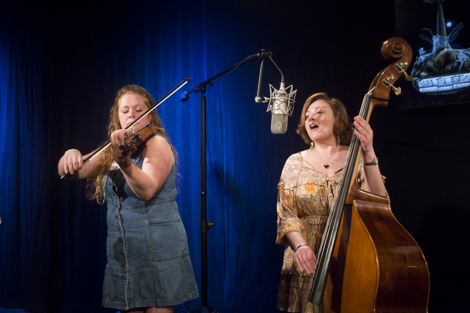 A woman plays violin while another woman playing upright bass sings into a microphone in a television studio.Katie Klan and Angela Brock of the Homer band Burnt Down House perform a Red Carpet Concert at KTOO Public Media during the 2019 Alaska Folk Festival. (Photo by Annie Bartholomew/KTOO)