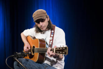 A man plays an acoustic guitar into a microphone on a blue background in a television studio. Guitarist Justin Smith performs a Red Carpet Concert at KTOO Public Media during the 2019 Alaska Folk Festival (Photo by Annie Bartholomew/KTOO)