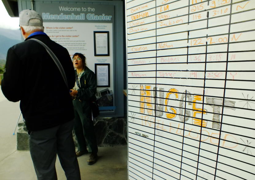 U.S. Forest Service interpreter Isabel Dzaik helps a tourist at the Mendenhall Glacier Visitor Center on July 14, 2019 as a nearby sign shows the closure of the Nugget Falls Trail because of a jökulhlaup or glacial dam release.