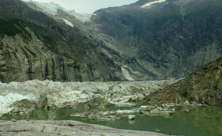 This is a wide view of Suicide Basin looking east from the edge of Mendenhall Glacier. A small, overhanging portion of the Juneau Icefield is visible in the upper left. Melt water from the icefield enters Suicide Basin from a large waterfall which is partially visible just below the ice. A portion of the ice dam, where most of the glacial dam release will occur, is visible at the lower left. Water spilling over the dam runs down to the right along the eastern edge of the glacier.
