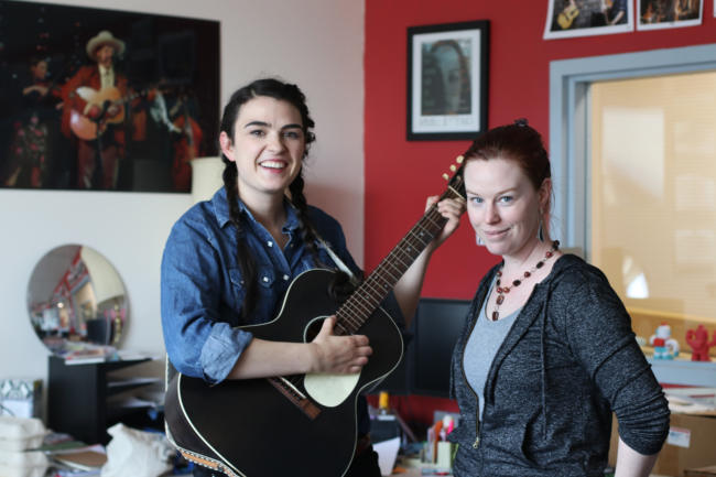 Two women with a guitar stand in front of a desk at KTOO Public Media. Juneau musicians Annie Bartholomew and Marian Call are performing at Friday's Souteast by Southwest: Music Showdown at the Gold Town Nickelodeon on August 2, 2019. Bartholomew is an Arts & Culture Producer at KTOO Public Media. (Photo by Stella Tallmon/KTOO Public Media)
