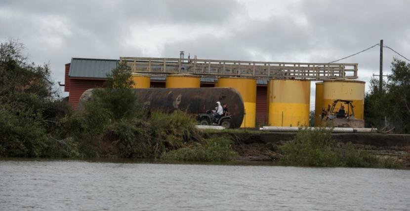 People ride on a four wheeler in front of the Napakiak school fuel tanks, which sit less than 100 feet from the Kuskokwim River following accelerating erosion.