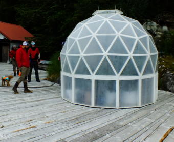 In this picture taken in early June 2019, Tom Lafollette, caretaker of the Annex Creek Hydroelectric Facility in Taku Inlet (obscured behind left side of greenhouse), explains to visitors how he built this scratch-built geodesic greenhouse for growing tomatoes, peppers, and other vegetables. The greenhouse is about 10 feet in diameter and is a slightly smaller version than a previous greenhouse he constructed from plans.