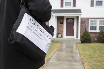 A man with a U.S. Census Bureau bag stands outside the front door of a suburban home.