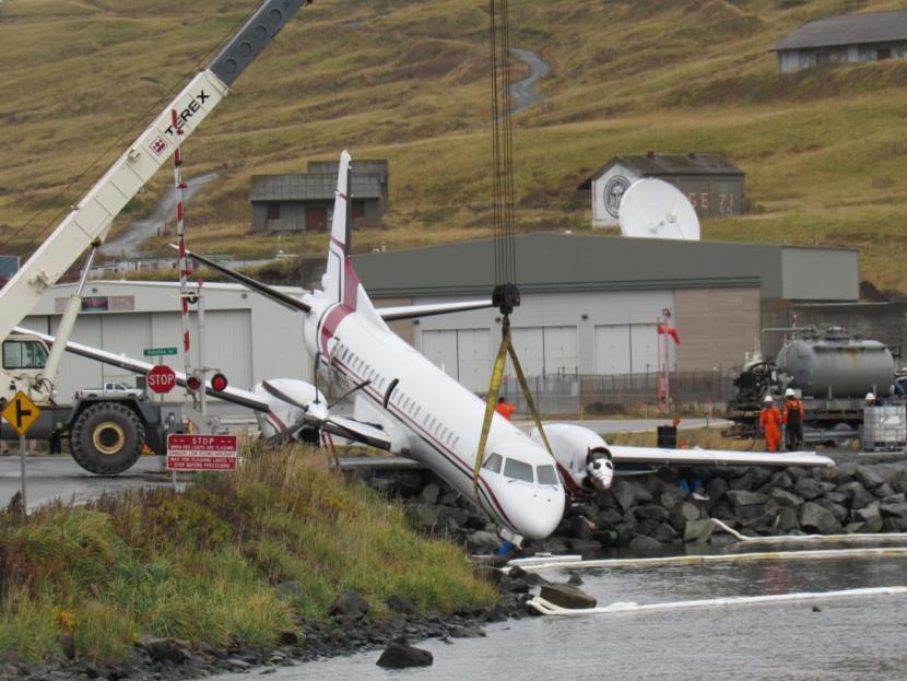 A crane lifts an airplane from a rocky drop near the runway.