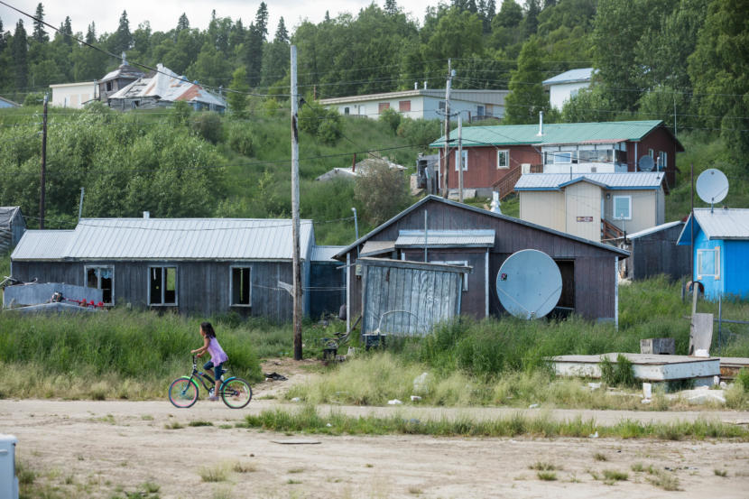 Syra Kozevnikoff, 9, rides her bicycle along a road on June 28, 2019 in Russian Mission. The tan building behind her is the village public safety office, which has three jail cells that are rarely used. (Photo by Loren Holmes/Anchorage Daily News)