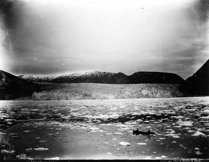 Two figures paddle a canoe amidst floating ice with Taku Glacier in background, circa 1895 (Alaska State Library - Historical Collections ASL-P87-1952)