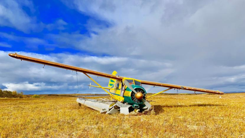 A small single-propeller floatplane sits in a yellow field, its landing gear crushed underneath.