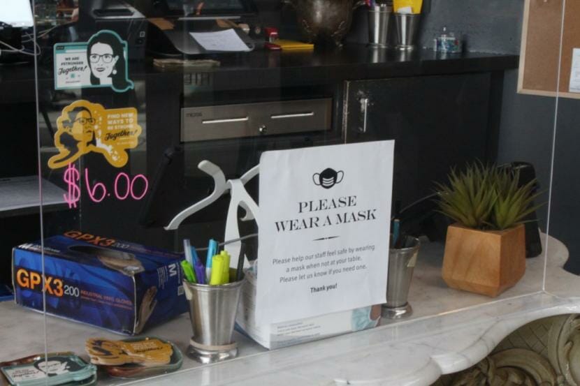 Inside a restaurant, a plexiglass shield, vinyl gloves, and sign asking patrons to wear masks.