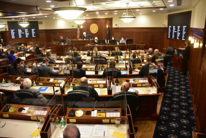 Alaska House representatives sit in their desks facing House Speaker Stutes. The back of their heads are facing the camera and plexiglass dividers are between each desk.